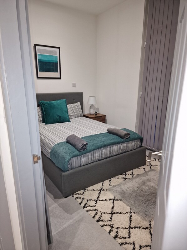 A bedroom with a bed, a nightstand, a lamp, a framed picture on the wall, and a patterned rug.