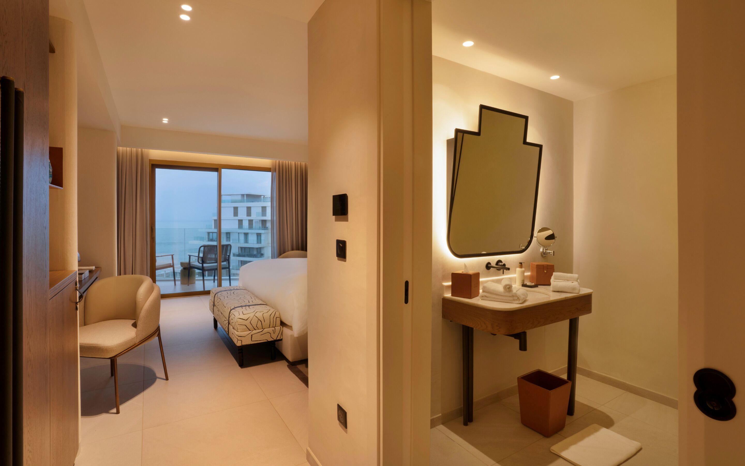 Une chambre d’hôtel avec un lit, une chaise, une petite table, une salle de bain avec un miroir et une vue sur les bâtiments par la fenêtre.