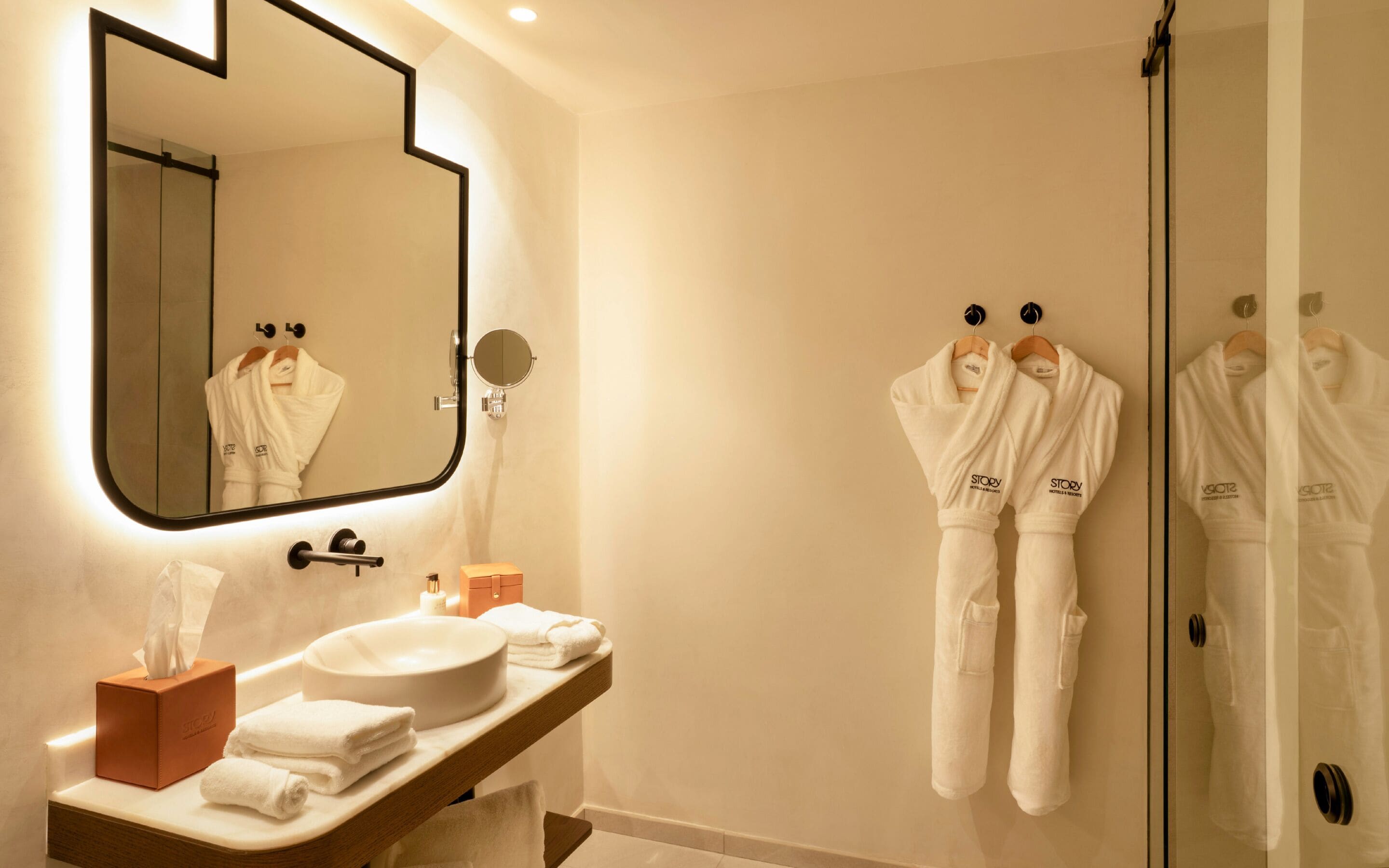 A modern bathroom with a large mirror, a round sink, and two white robes hanging on hooks.