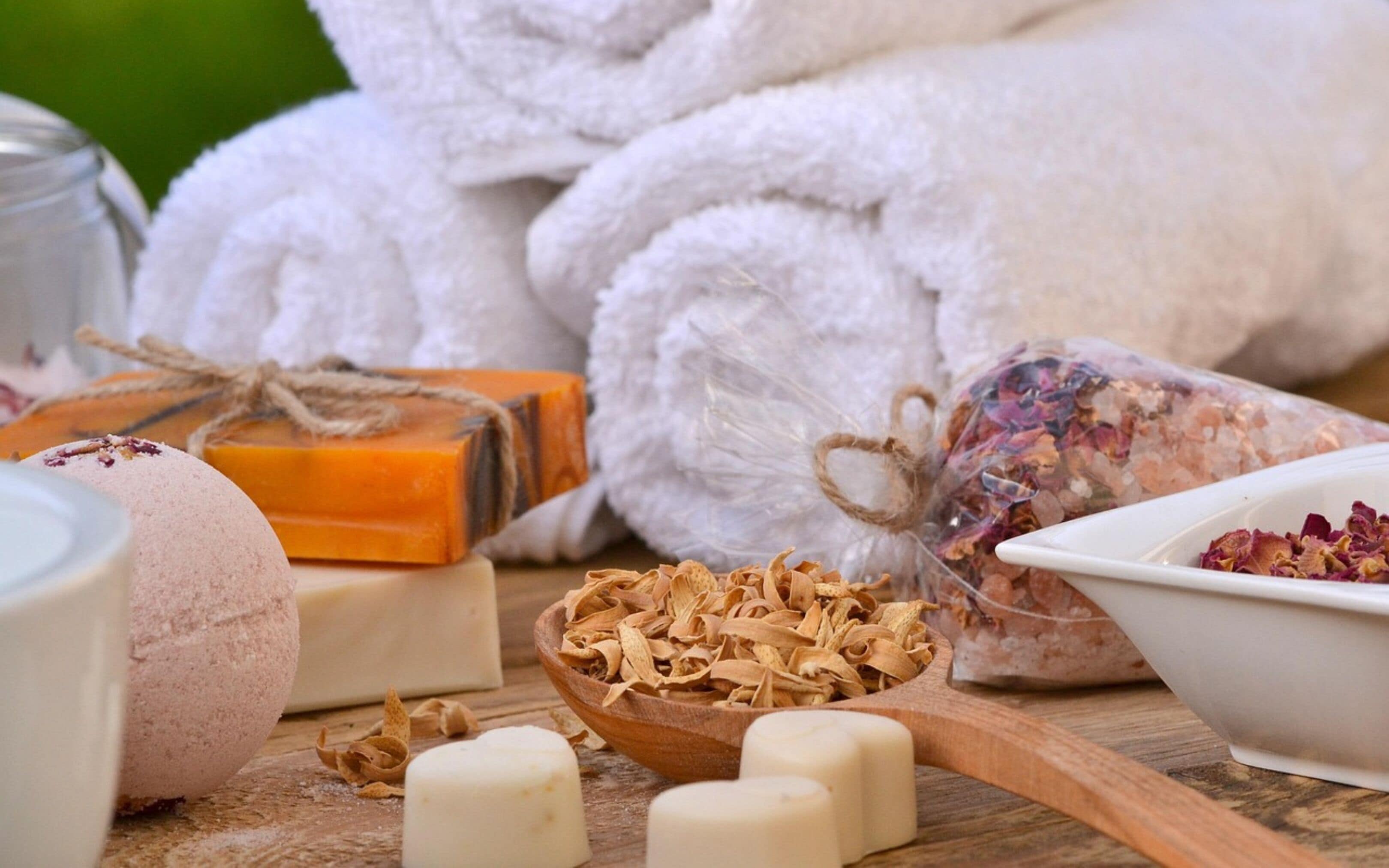A wooden table with various bath products including soaps, a wooden spoon with dried flowers, and a clear bag with dried petals.
