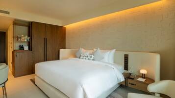 A modern hotel room with a large bed, a wooden wardrobe, a bedside table with a lamp, and a small seating area.