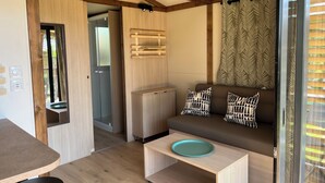 Un espace de vie compact doté d’un plafond en bois, d’un canapé avec des coussins à motifs, d’une petite table avec une assiette, et d’une porte-fenêtre coulissante donnant sur un espace extérieur.