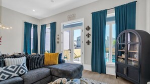 Een woonkamer met een grijze hoekbank, turquoise gordijnen, een zwart kastje en een glazen salontafel.