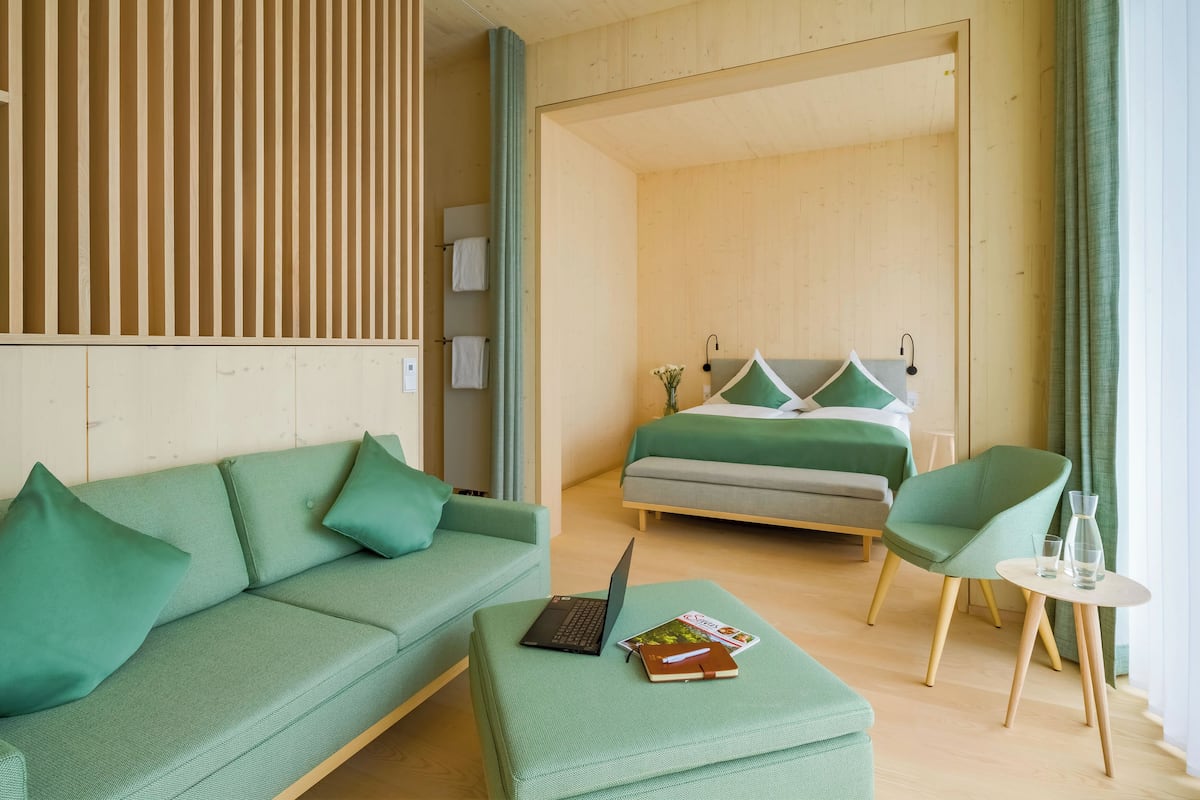 A modern hotel room with a green sofa, a small table with a laptop and a book, a green armchair, and a bed with green bedding.