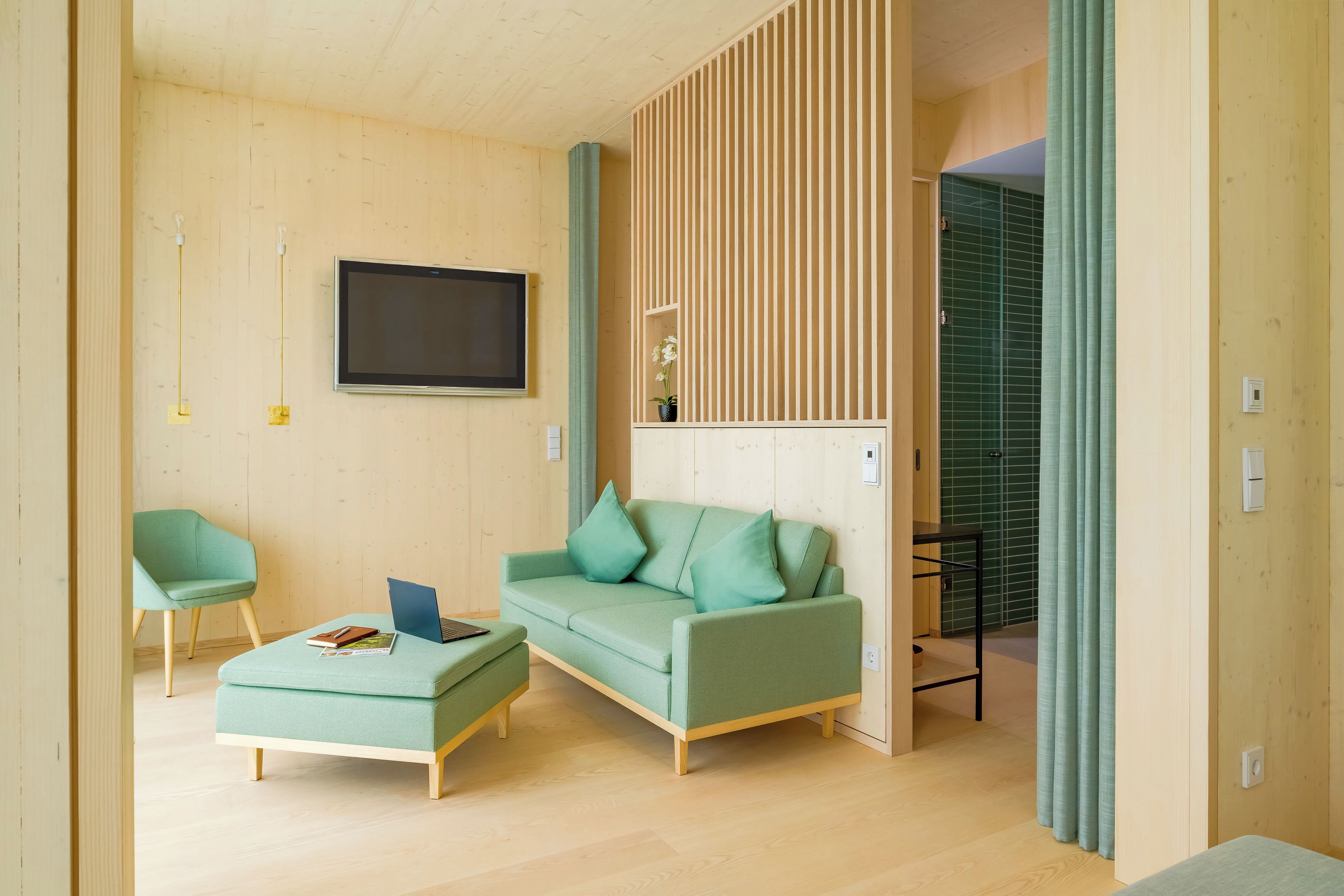 A modern living room with a green sofa, a matching ottoman, and a teal armchair. A flat-screen TV is mounted on the wall, and there is a small table with a laptop and books.