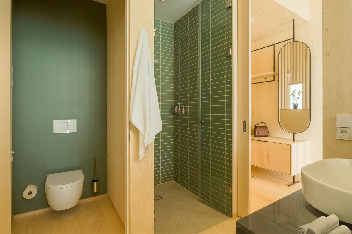 A bathroom with a toilet, green tiled shower, wooden vanity, and a large mirror.