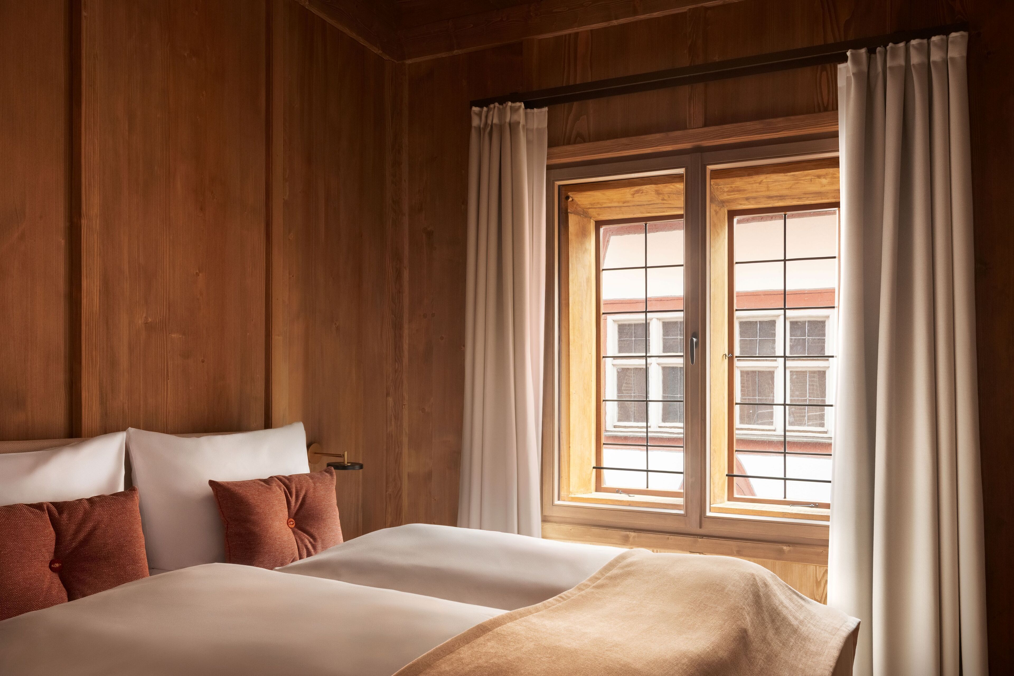 A room with wooden panel walls, a large window with white curtains, and a bed with white and brown bedding.