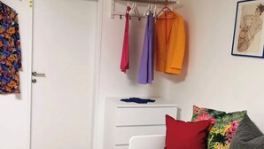 Een slaapkamer met een witte kast, een bed met kleurrijke kussens en een kledingrek met verschillende kledingstukken.
