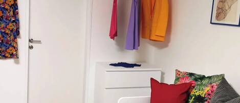 A bedroom with a white wardrobe, a bed with colorful pillows, and a clothing rack with various garments.