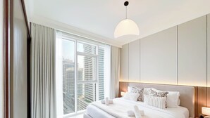 Une chambre d’hôtel moderne avec un grand lit, une table de chevet avec une lampe, une vue sur de hauts immeubles à travers de grandes fenêtres et un luminaire au plafond.