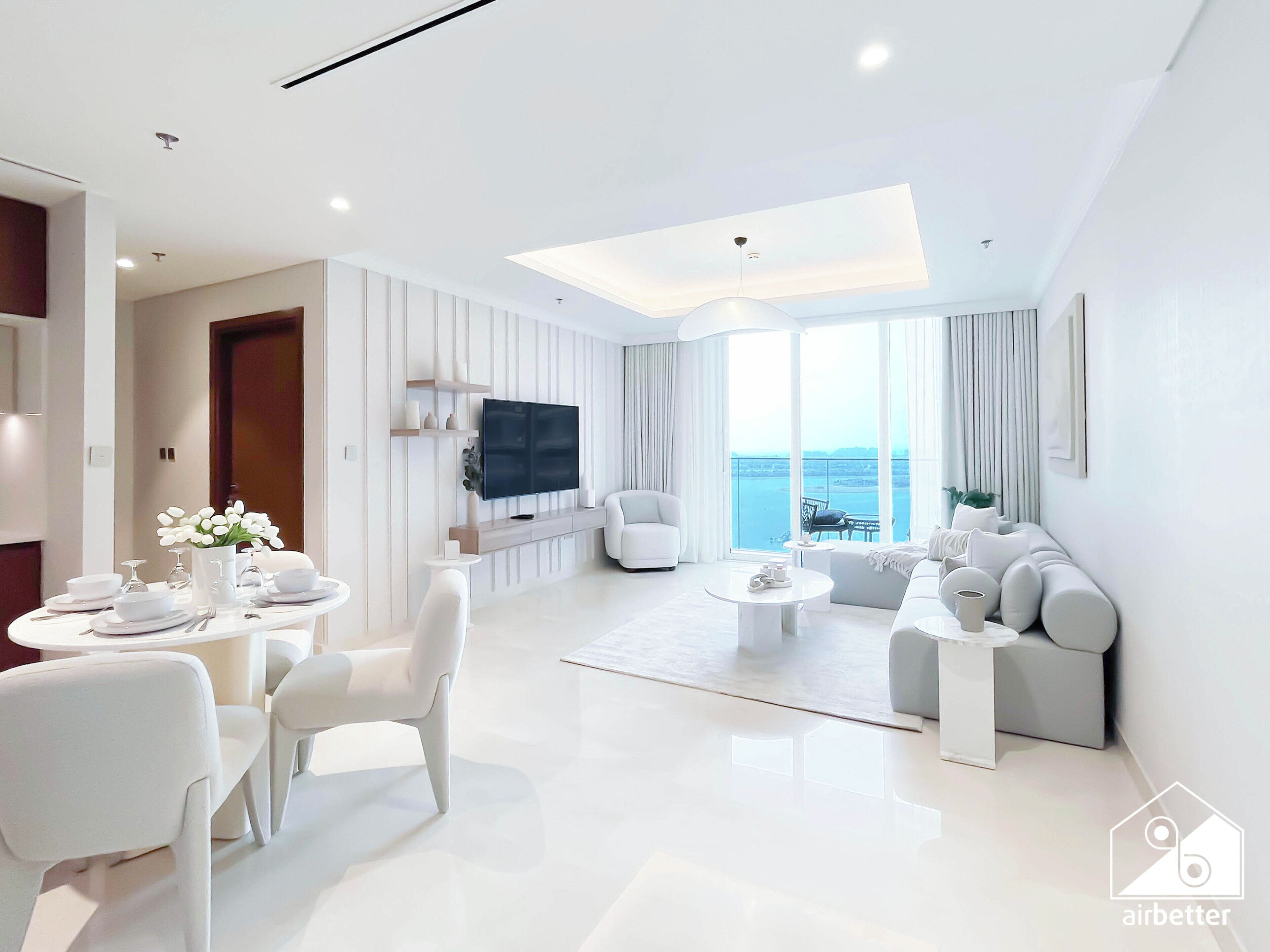 Un soggiorno moderno con una televisione a schermo piatto, arredi bianchi, un'area pranzo con tavolo imbandito per quattro persone e un'ampia finestra con vista sul mare.