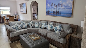 Un salon avec un canapé d’angle gris, un pouf gris et un tableau représentant une scène de plage.