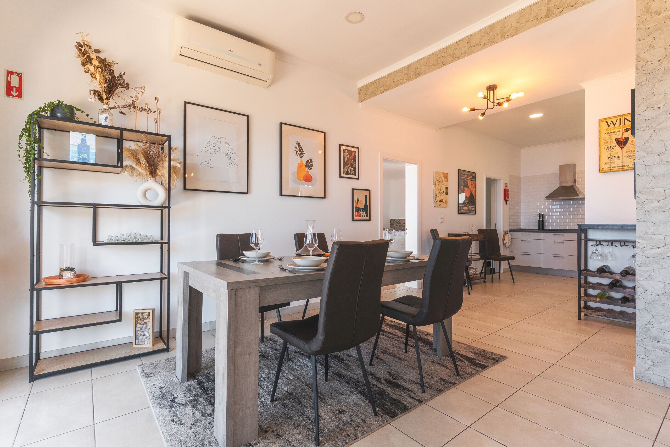A modern dining area with a wooden table, black chairs, and a decorative shelf with plants and framed pictures.