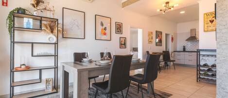 A modern dining area with a wooden table, black chairs, and a decorative shelf with plants and framed pictures.