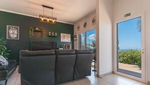 Een moderne woonkamer met een donkergroene wand, een zwarte leren bank, een flatscreen televisie en een grote schuifglazen deur die uitkomt op een balkon met uitzicht op de oceaan.
