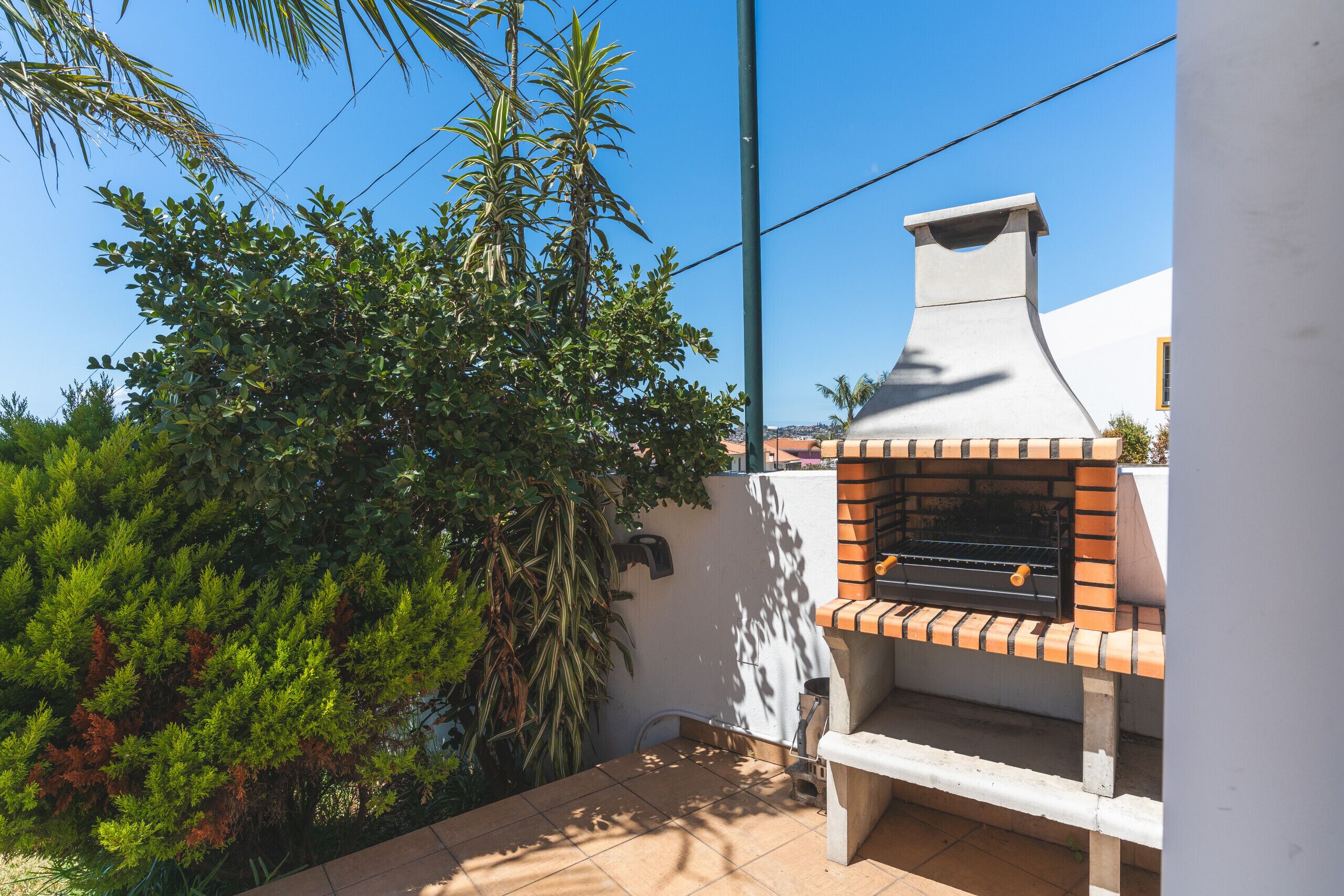 A patio area with a brick oven grill, surrounded by greenery and a white wall.