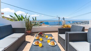 Een terras op het dak met rieten meubilair, ontbijt met fruit en croissants, en uitzicht op zee.