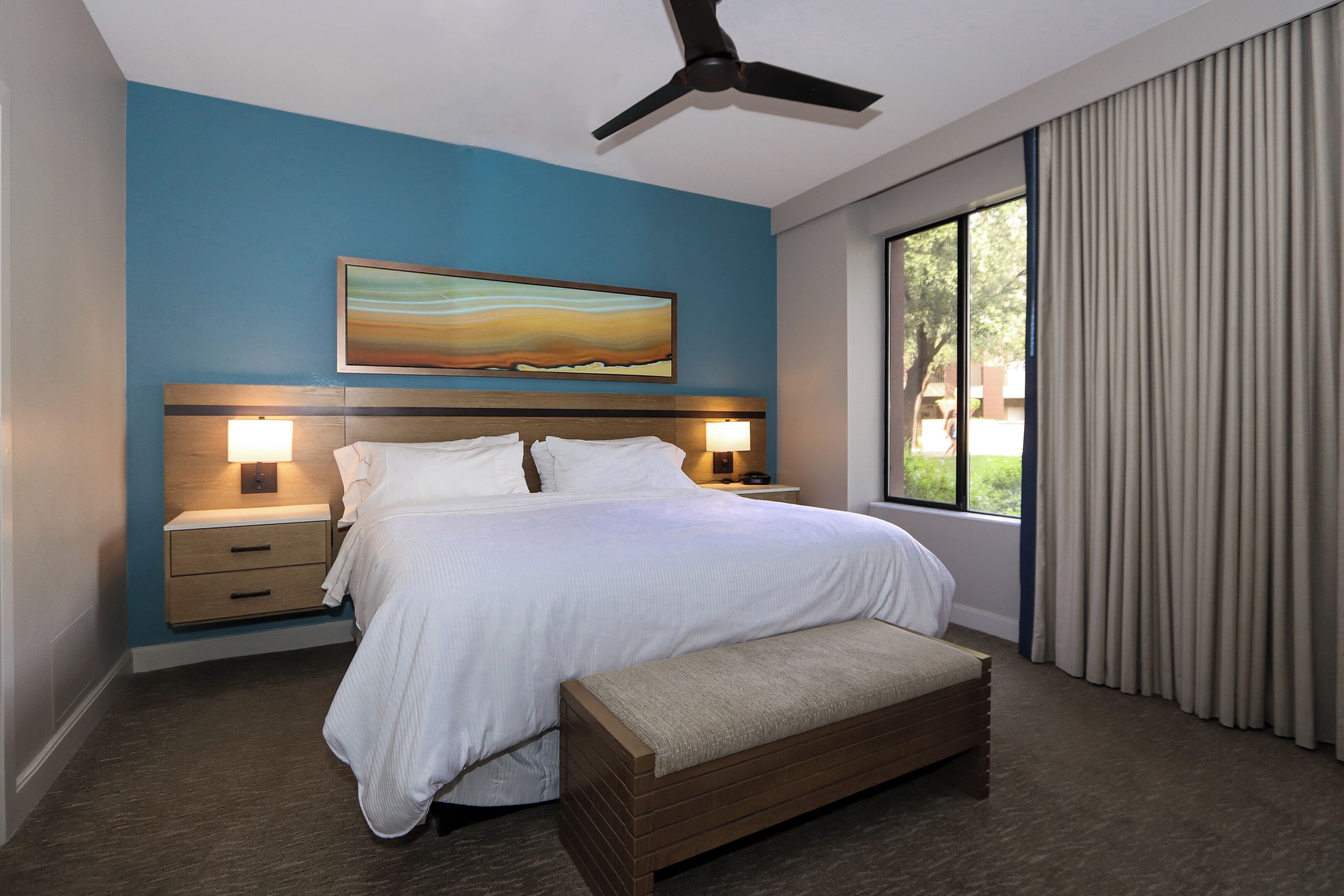 Een moderne hotelkamer met een groot bed, bedlampjes, een plafondventilator, een raam met gordijnen en een schilderij aan de muur.