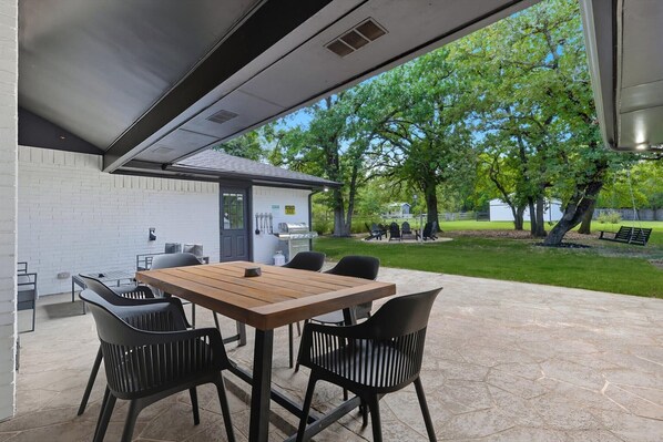 An outdoor patio with a wooden table and black chairs, a grill, and a white brick wall.