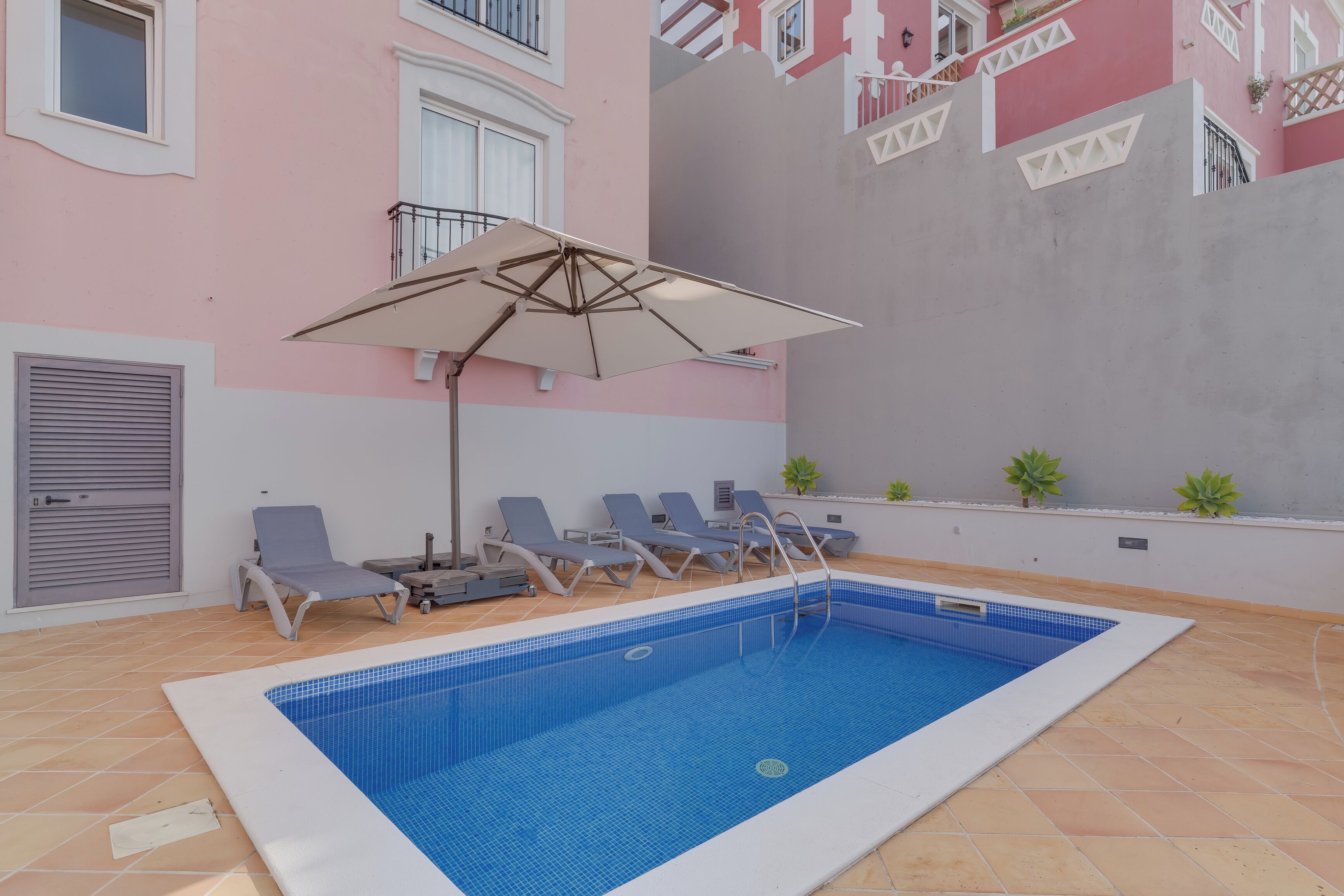 A swimming pool with blue tiles, surrounded by lounge chairs and a large umbrella.