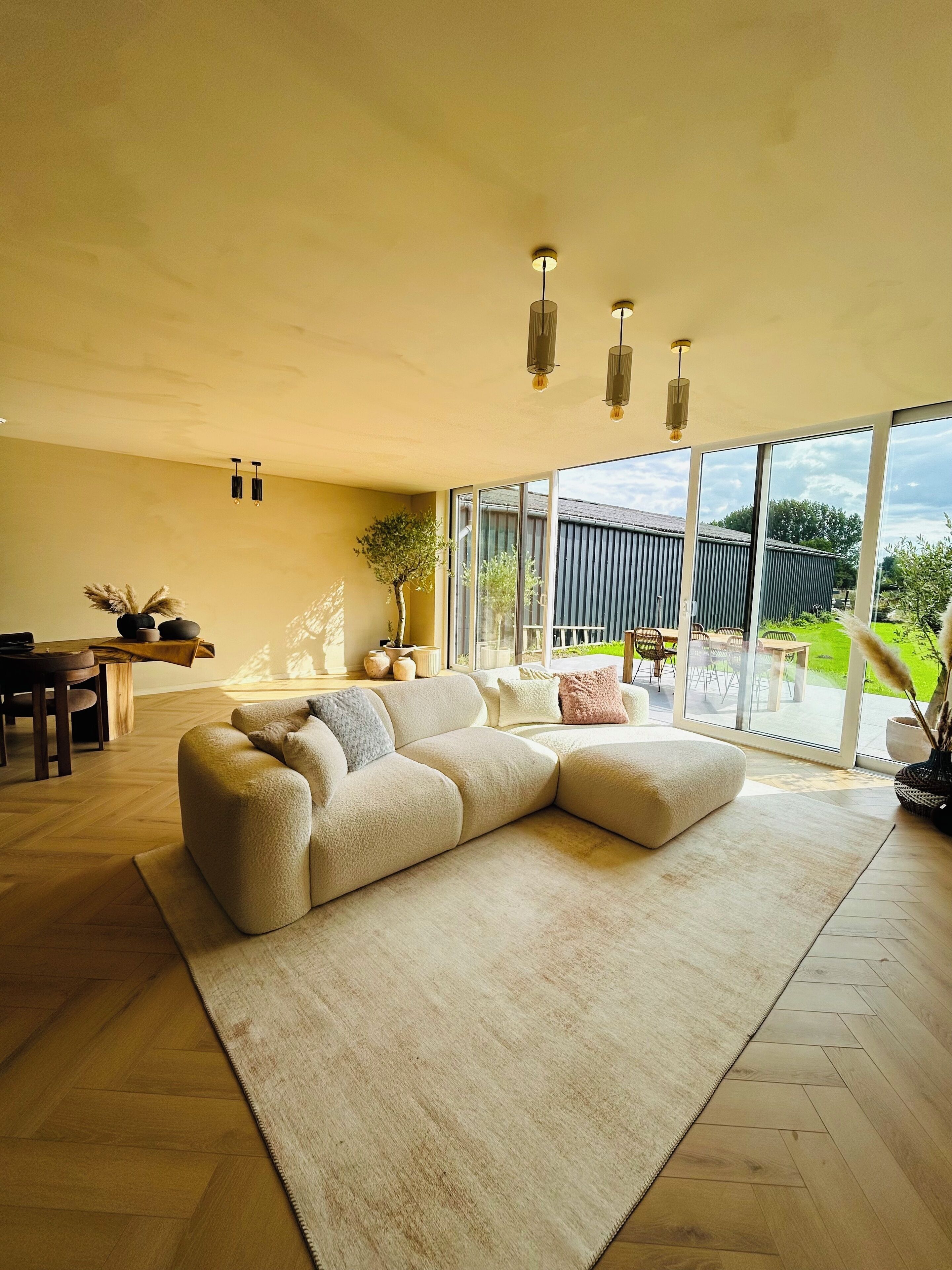 Un salon spacieux avec un grand canapé d’angle, un coin repas doté d’une table en bois et de grandes fenêtres offrant une vue sur l’extérieur.