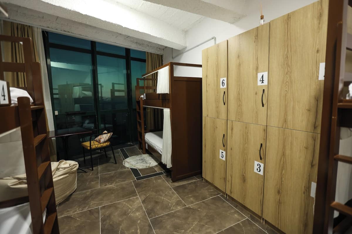 A dormitory room with bunk beds, a desk, and a chair. There are wooden cabinets with numbered compartments.