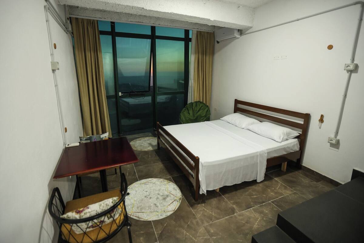 A small, clean room with a bed, a desk, and a chair. There is a window with curtains, a small table, and a round rug.