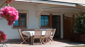 A covered outdoor seating area with a table and chairs, a wooden door, and a window with a blue patterned curtain. - THE LADY OF THE LAKE (Molveno)