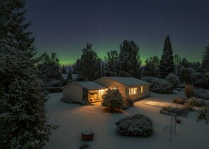 Une maison dont la cour est recouverte de neige, entourée d’arbres à feuilles persistantes, sous un ciel étoilé.