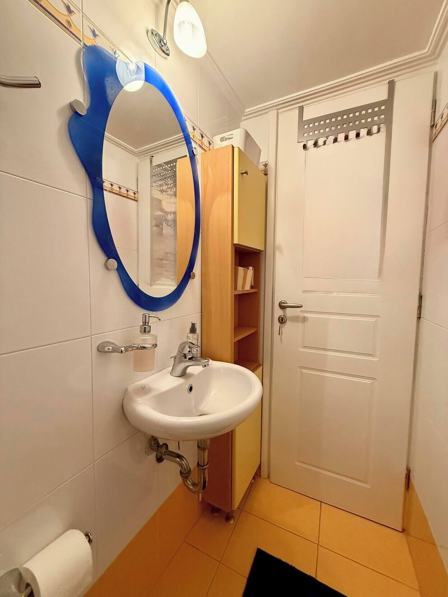 A bathroom with a white sink, a mirror with a blue frame, a towel dispenser, and a wooden cabinet.