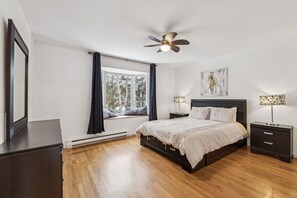 A bedroom with a bed, a ceiling fan, a window with curtains, a nightstand, and a wall-mounted artwork.