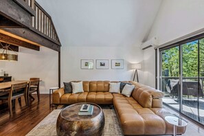 A modern living room with a large sectional sofa, a wooden dining table, and a large window with a view of the outdoors. - Luxury Snow Village Ski-In/Out, Golf & Club Access (Hartford)