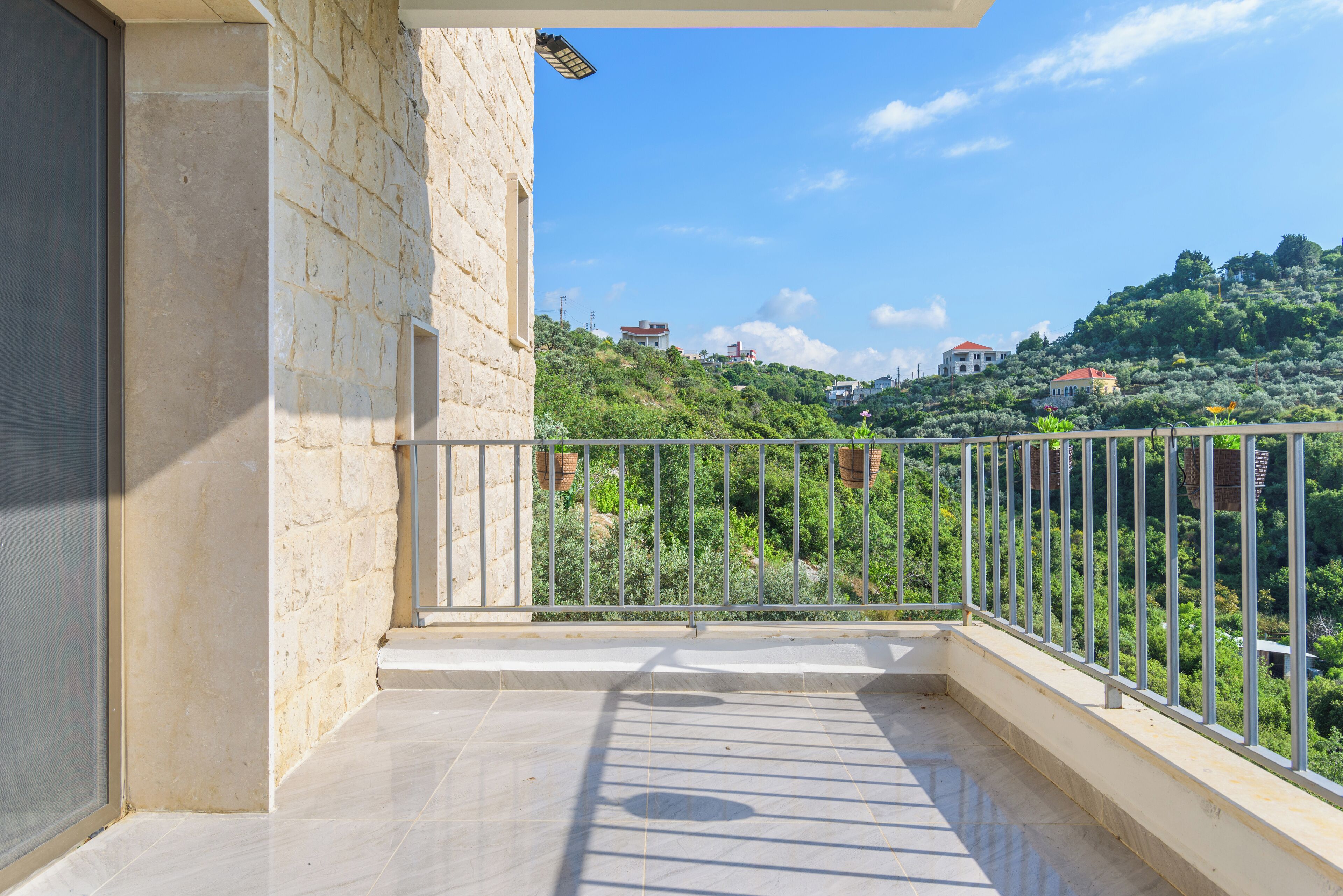 A balcony with a stone wall and metal railing, overlooking a hillside with buildings and greenery.
