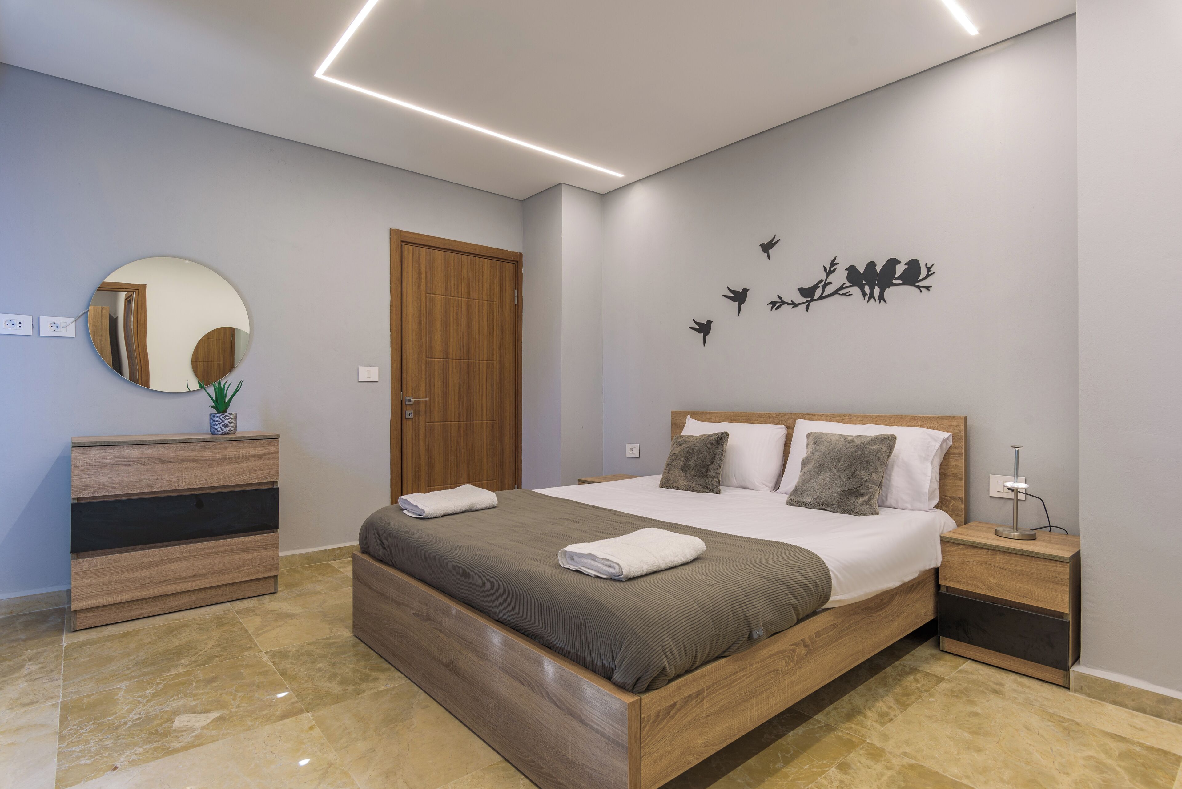 A modern bedroom with a wooden bed, a nightstand, a dresser, a mirror, and a wall art of birds on a branch.