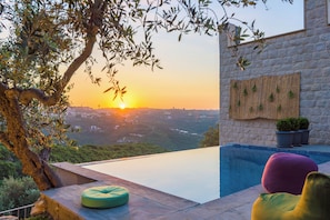 Une piscine à débordement offrant une vue sur le coucher de soleil au-dessus d’une vallée, un mur de pierre avec un panneau en bois et un arbre aux branches pendantes.