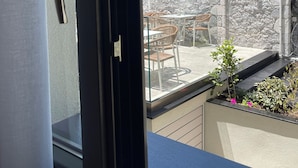 A view through a window of a balcony with outdoor seating, a stone wall, and a pink building in the background. - Akti Vigla Premium (Lesvos)