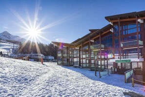 Un centro de esquí cubierto de nieve con un albergue de varios pisos, balcones y grandes ventanales.