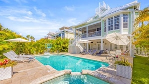 A swimming pool with lounge chairs and umbrellas, surrounded by a house with a balcony and a view of palm trees. - Summer Breeze 7 Bedroom Home (Captiva)