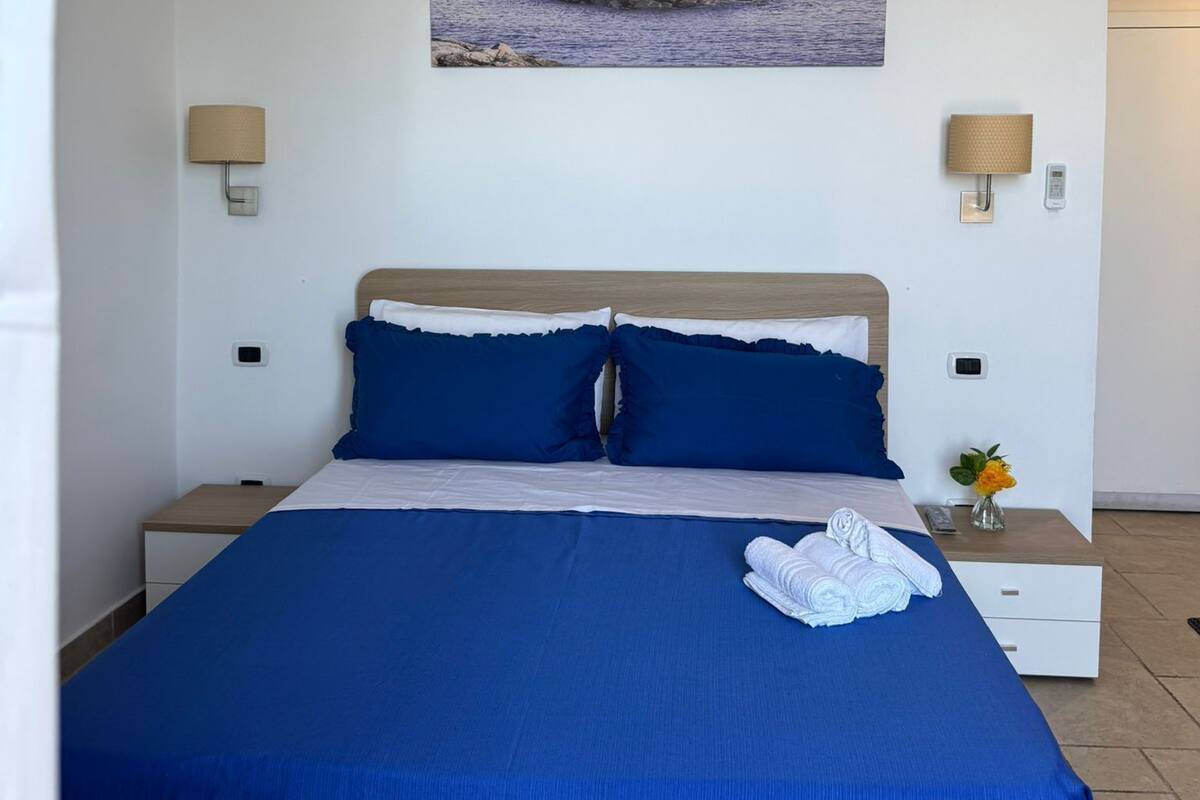 A bedroom with a bed, two bedside lamps, a nightstand, and a painting of a coastal scene.