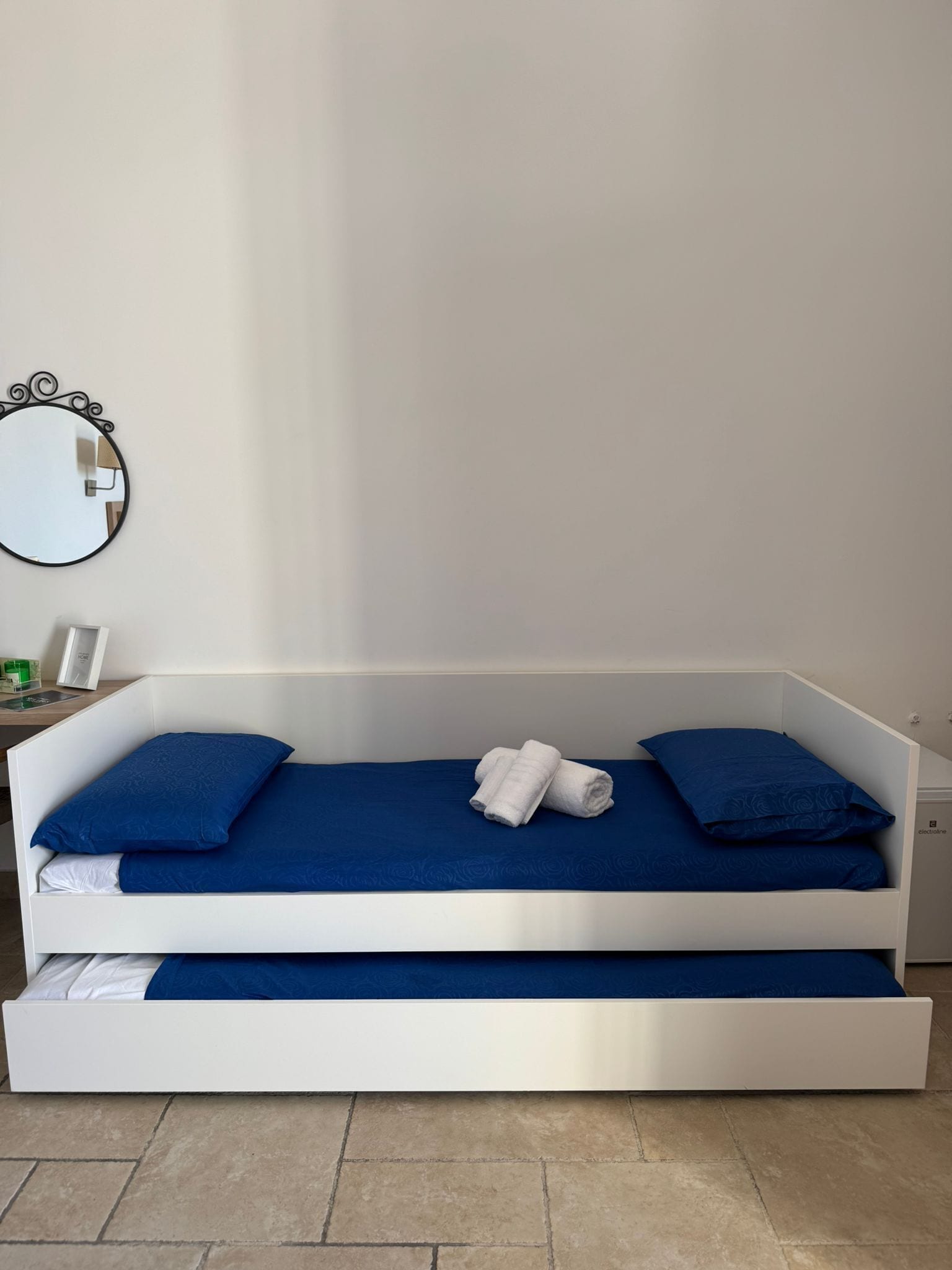A white bed with blue bedding and pillows, a small bedside table with a lamp, and a round mirror on the wall.