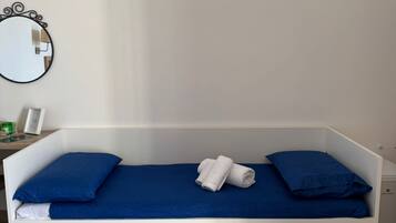 A white bed with blue bedding and pillows, a small bedside table with a lamp, and a round mirror on the wall.