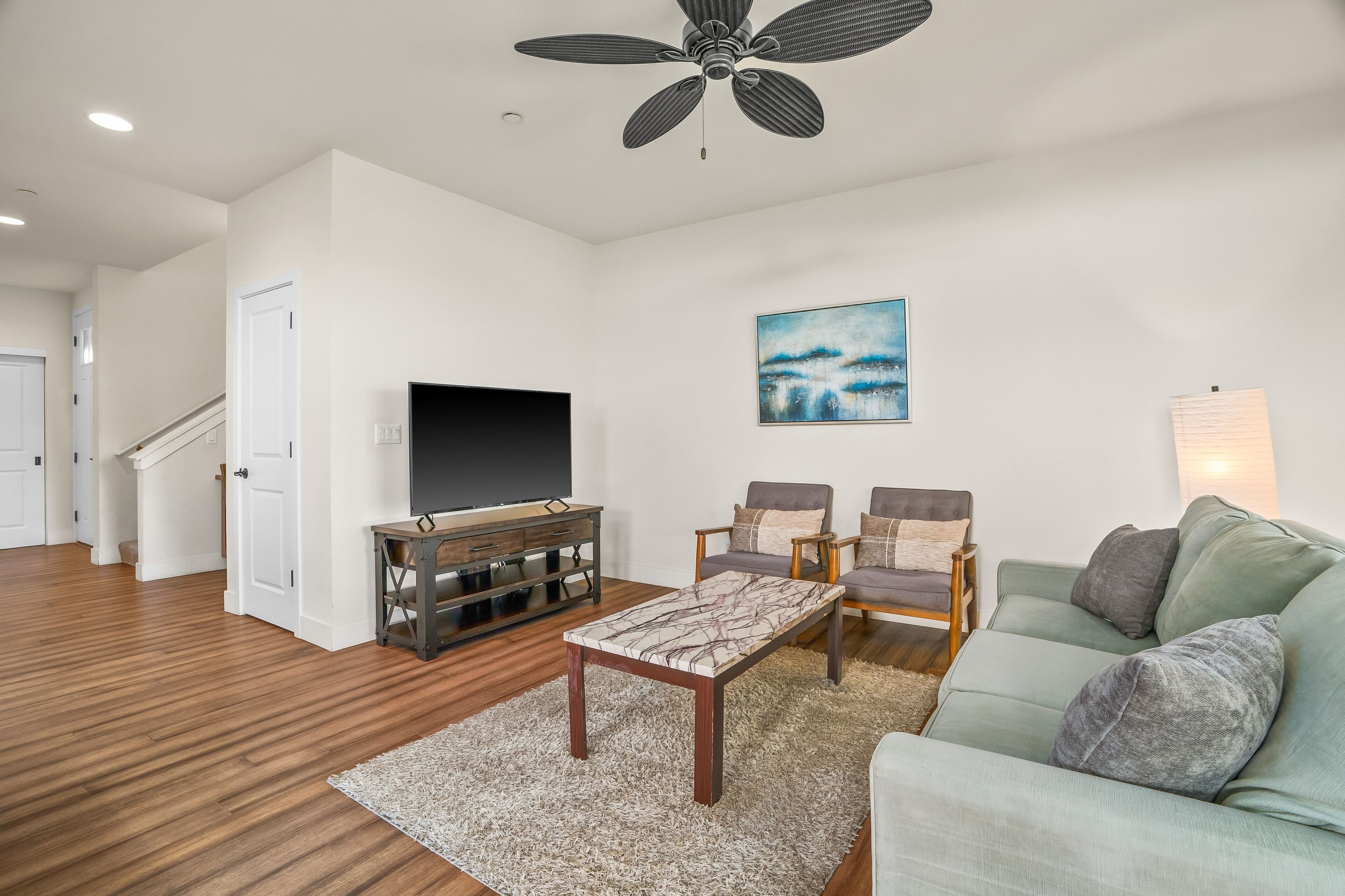 A living room with a flat-screen TV, two armchairs, a sofa, a coffee table, and a ceiling fan.