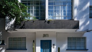 Un bâtiment avec une porte bleue portant l’inscription « JB71 », deux cactus en pot et un balcon orné de plantes.