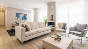 Een moderne woonkamer met een bank, fauteuils en een salontafel. Er is een open haard, een eethoek en een groot raam.