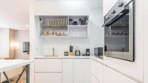 Een moderne keuken met witte kasten, een ingebouwde oven en een magnetron. Boven de aanrecht staan planken met servies en bestek.