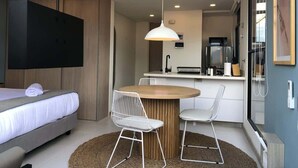 A modern hotel room with a bed, a dining area with a table and chairs, a kitchen area, and a large window.