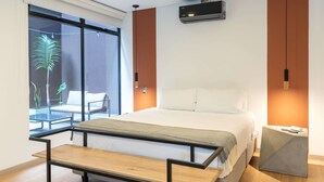Ein modernes Schlafzimmer mit einem großen Bett, einer Bank, einem Glastisch und Blick auf einen Balkon mit Pflanzen.