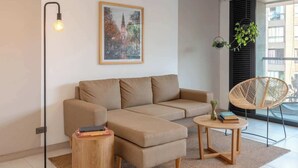 Un salon moderne avec un canapé d’angle beige, une table basse ronde en bois, un lampadaire et un fauteuil en osier.