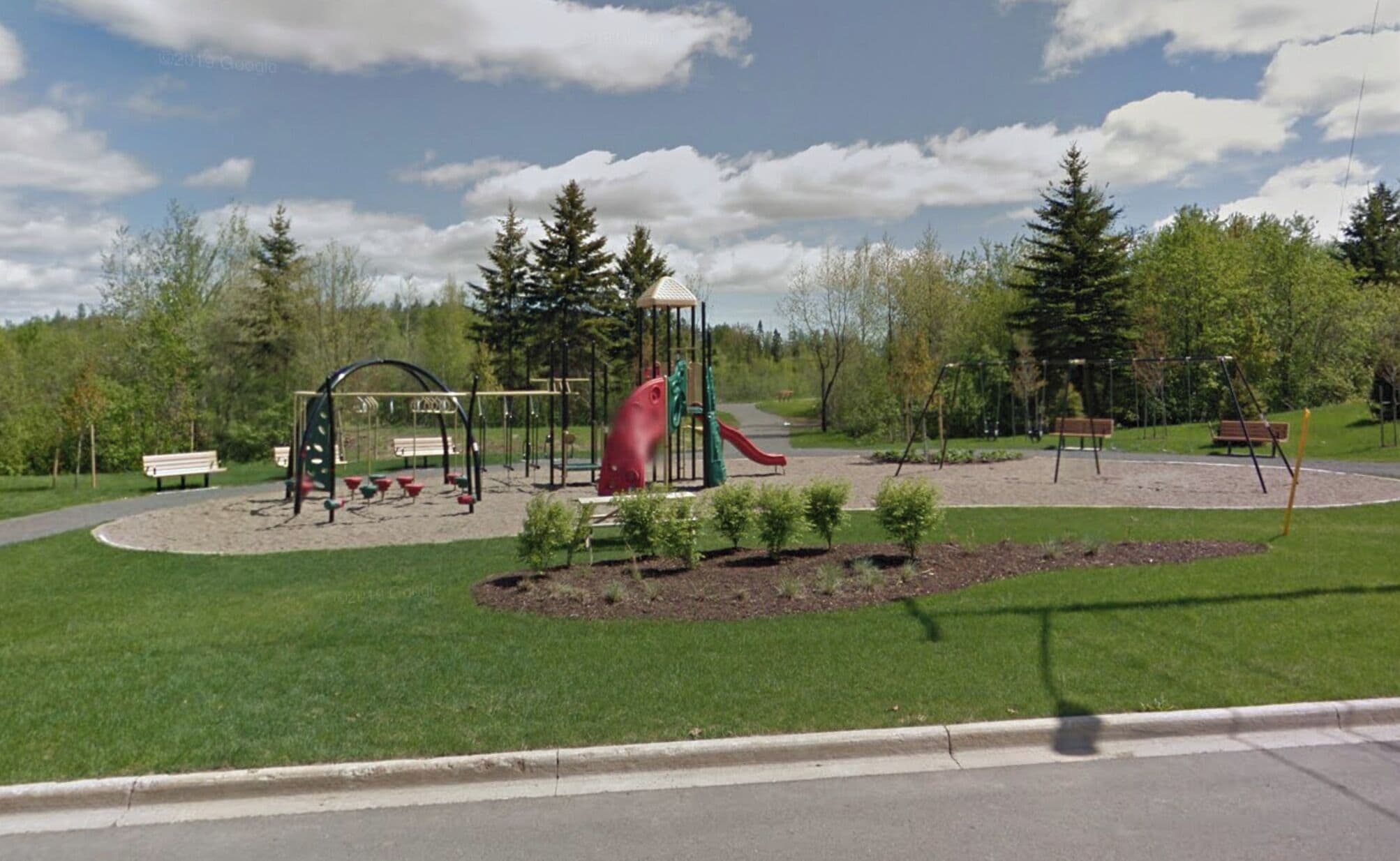 Ein Spielplatz mit Schaukeln, einer Rutsche und einer Kletterkonstruktion, umgeben von Gras und Bäumen.