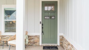 A green door with a 'WELCOME' sign, a stone wall, and a wicker chair outside.